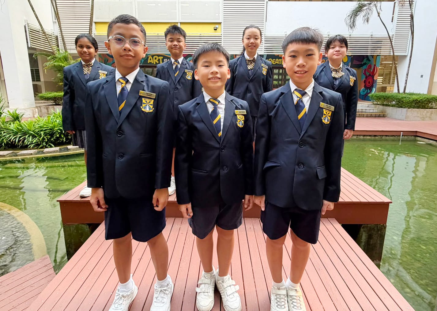 Seven students in uniforms stand before a Beacon Primary School wall showing mission, vision, and speech bubbles.